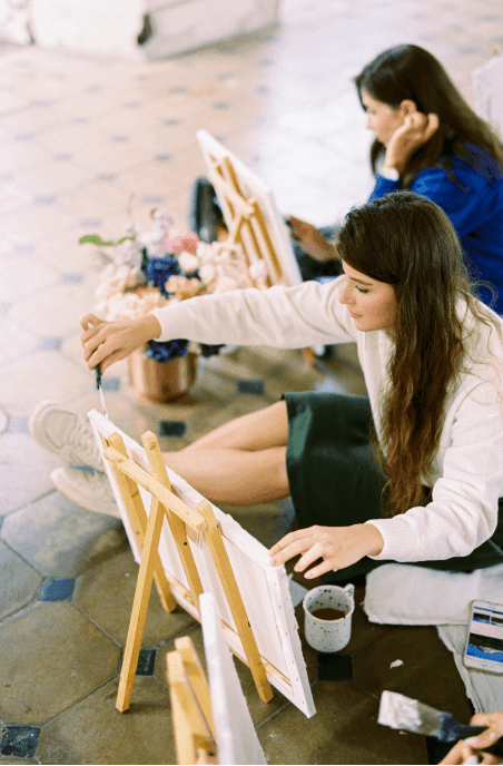 Two women sitting on the floor painting on canvases with art supplies and floral arrangement nearby.